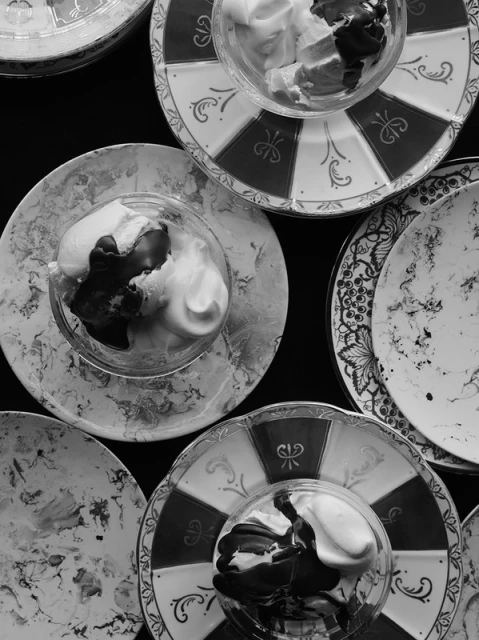 Monochrome photograph showcases glasses filled with ice cream on decorative plates, arranged on a dark surface, shot from above.