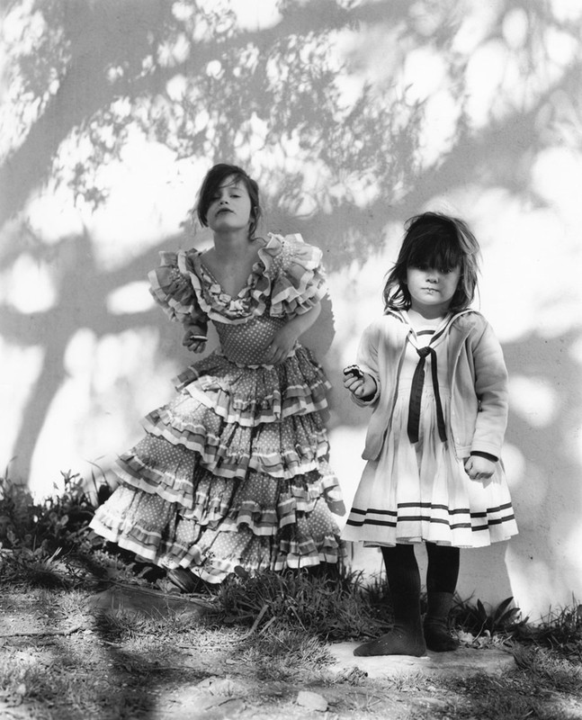 Two children pose outside in a vintage black and white photograph. One girl wears a tiered ruffled dress, the other in a sailor dress.