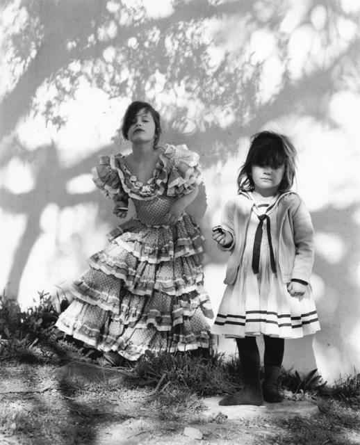 Two children pose outside in a vintage black and white photograph. One girl wears a tiered ruffled dress, the other in a sailor dress.