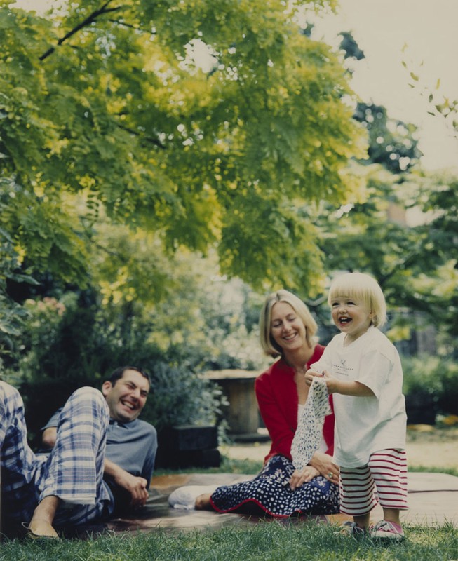 A family of three—a dad, mom, and toddler—laughing in a backyard garden under a tree, sitting on the grass in casual clothes on a sunny day.