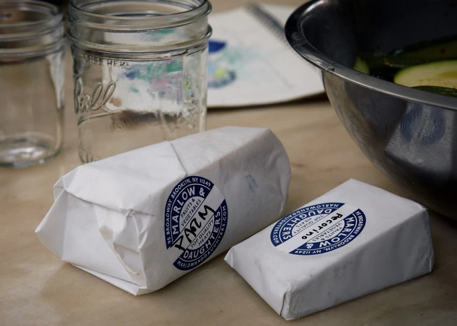 Wrapped Marlow & Daughters Farm goods sit near glass jars and a metal bowl of zucchini on a table, evoking a fresh, local food theme.