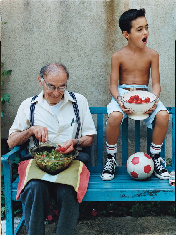 Elderly man and young boy sit together on a blue bench outdoors. The man prepares a salad, while the boy holds a bowl of tomatoes.