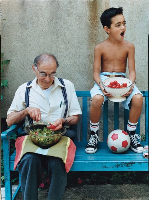 Elderly man and young boy sit together on a blue bench outdoors. The man prepares a salad, while the boy holds a bowl of tomatoes.