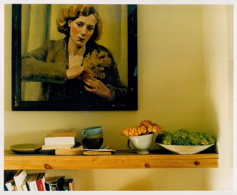 Still life of a portrait painting above a wooden shelf with books, flowers, cups, and a dish against a neutral colored wall in an interior space.