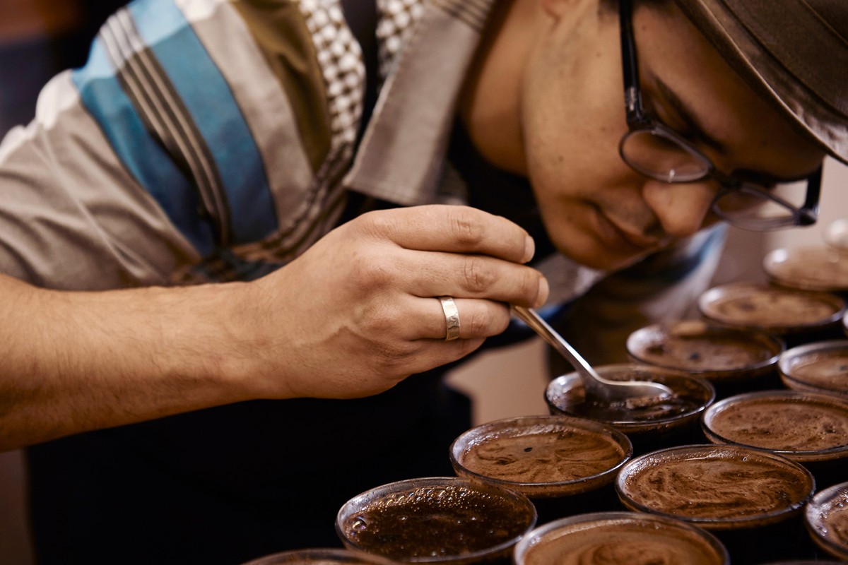 Close up of a man in eyeglasses using a spoon to sample liquid from multiple cups in a dark room. Coffee and food industry theme.