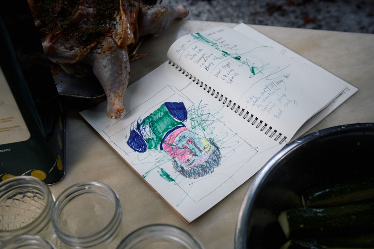 A close-up shot reveals an open sketchbook displaying colorful scribbled art, alongside a cooked bird, bowl of zucchini, and jars.
