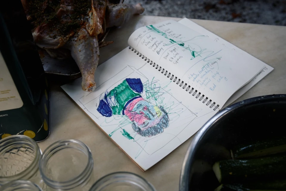 A close-up shot reveals an open sketchbook displaying colorful scribbled art, alongside a cooked bird, bowl of zucchini, and jars.