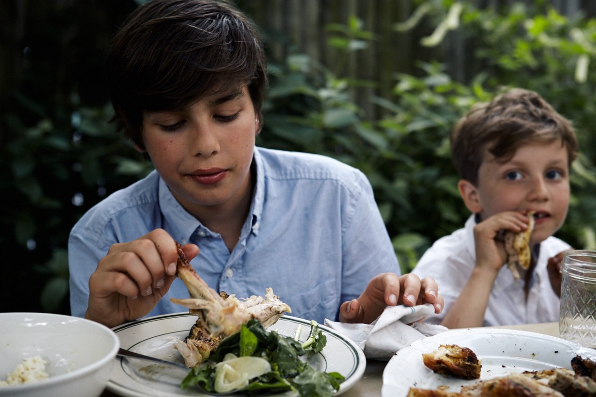 Two boys eat chicken and salad at an outdoor table surrounded by green foliage, enjoying a meal together in a natural setting.
