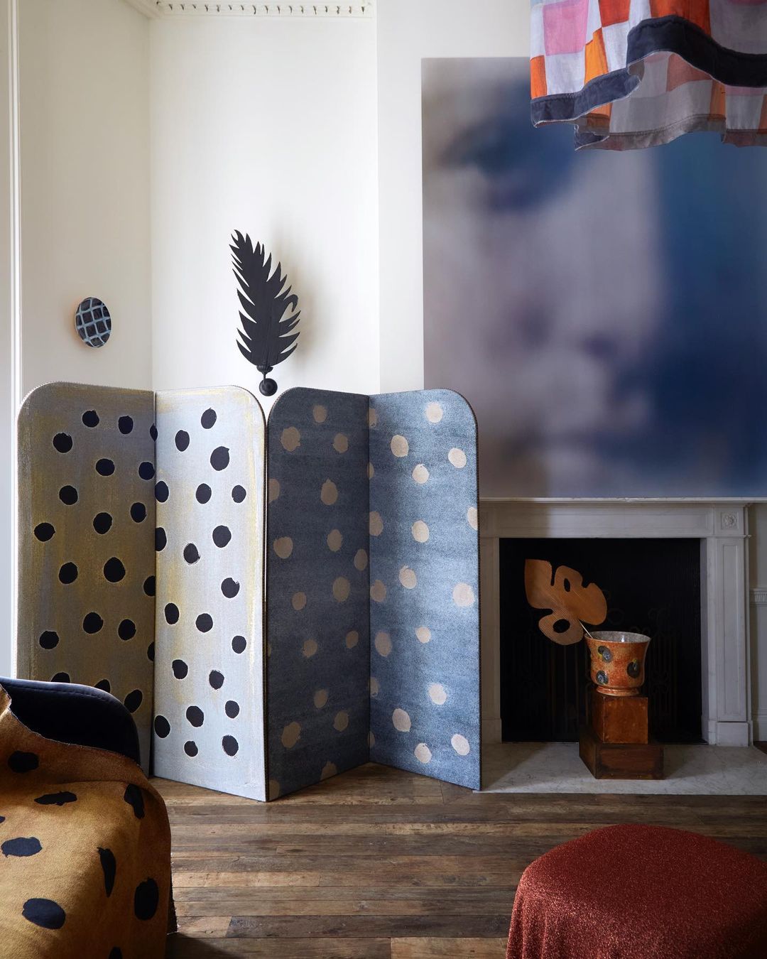 Interior of a room featuring a folding screen with polka dots, a modern painting above a fireplace, and a patterned armchair and ottoman.