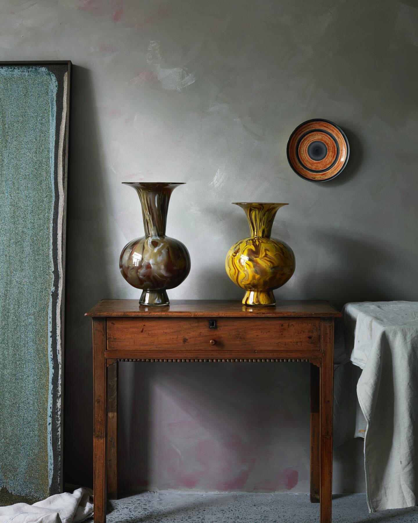 Two vases sit on a wooden table against a grey wall with abstract art and a decorative plate, showcasing home decor and interior design.