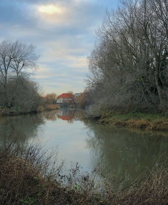 Calm river reflecting bare trees and distant buildings under an overcast sky by photographer Martyn Thompson