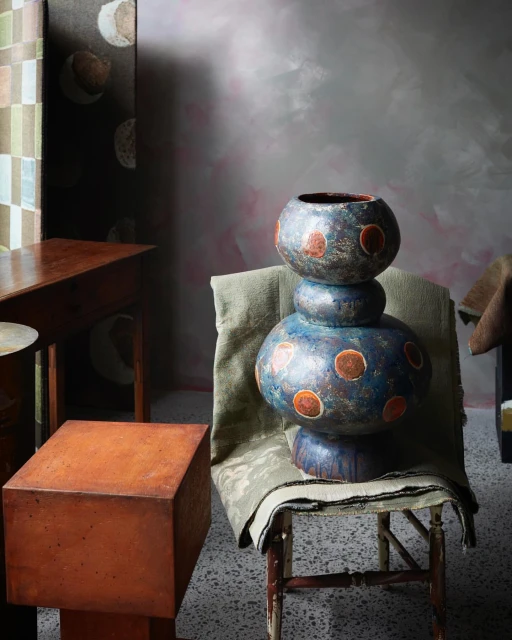 Elevated view of a tall, handmade ceramic vase on a wooden chair with patterned fabrics, set against a textured grey-pink wall.