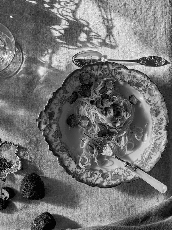 Overhead shot of truffle pasta in ornate bowl, with truffle shavings, glass, spoon, fork, and leaf shadows on a white linen surface.