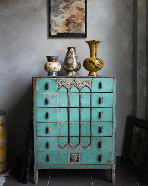 Antique turquoise chest of drawers against a grey wall, topped with three decorative vases and a framed picture above.