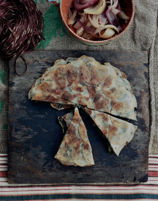 Overhead shot of a savory pie cut open on a rustic wooden board, with onions in a bowl and strands of rope nearby.