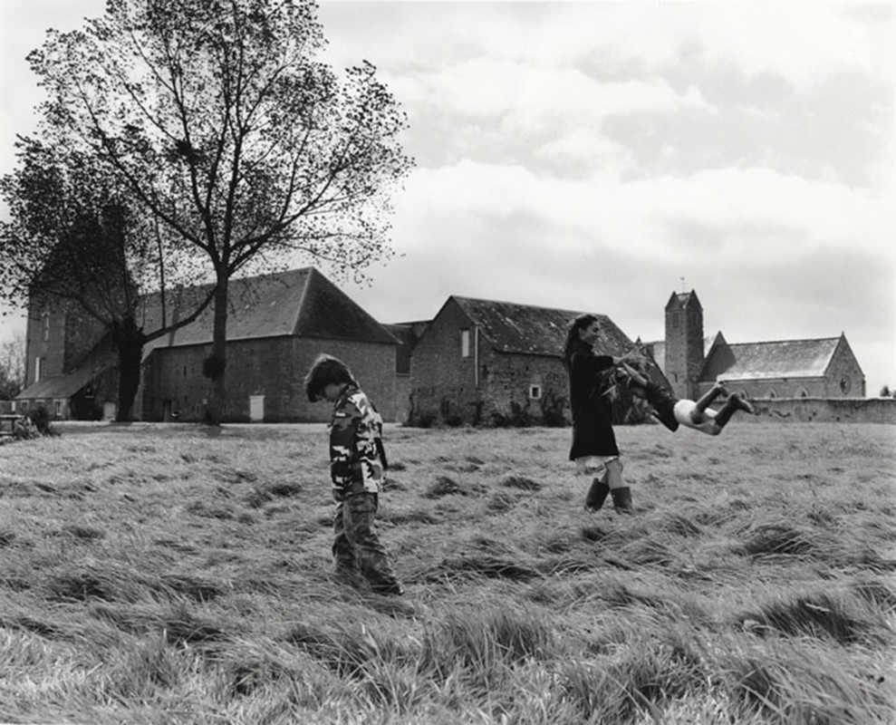 Black-and-white image showing child in camouflage walking on grass, woman tossing child, building and tree in background.