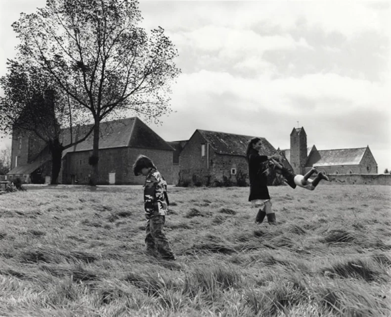 Black-and-white image showing child in camouflage walking on grass, woman tossing child, building and tree in background.