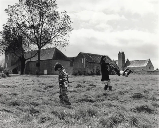 Black-and-white image showing child in camouflage walking on grass, woman tossing child, building and tree in background.