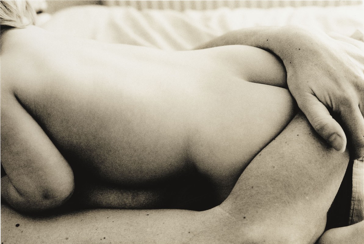Close-up of a nude adult female reclining on a bed with an arm across her, in sepia tones, with a shallow depth of field.