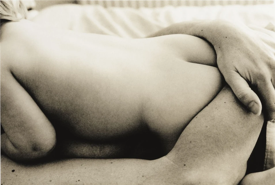 Close-up of a nude adult female reclining on a bed with an arm across her, in sepia tones, with a shallow depth of field.