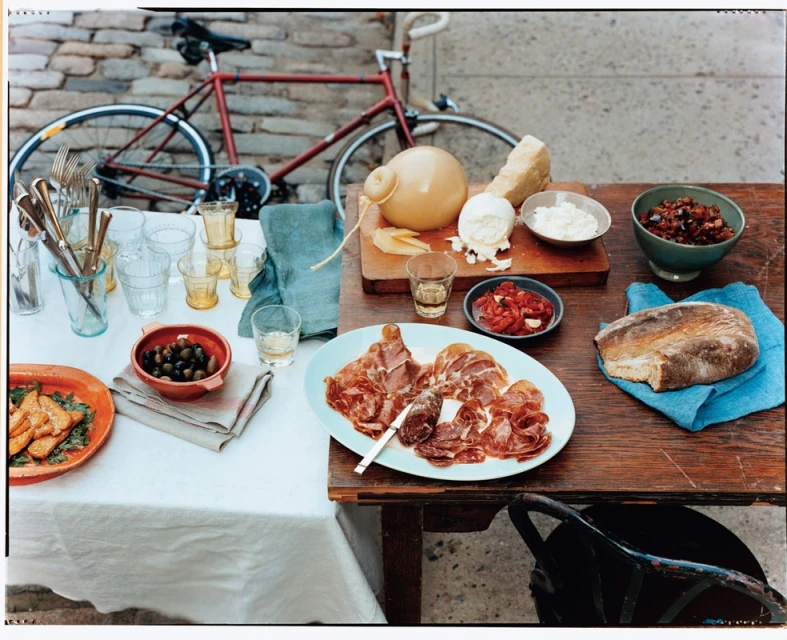 Rustic food display on a wooden table with Italian cheeses, cured meats, bread, olives, and a red bicycle parked behind outdoors.