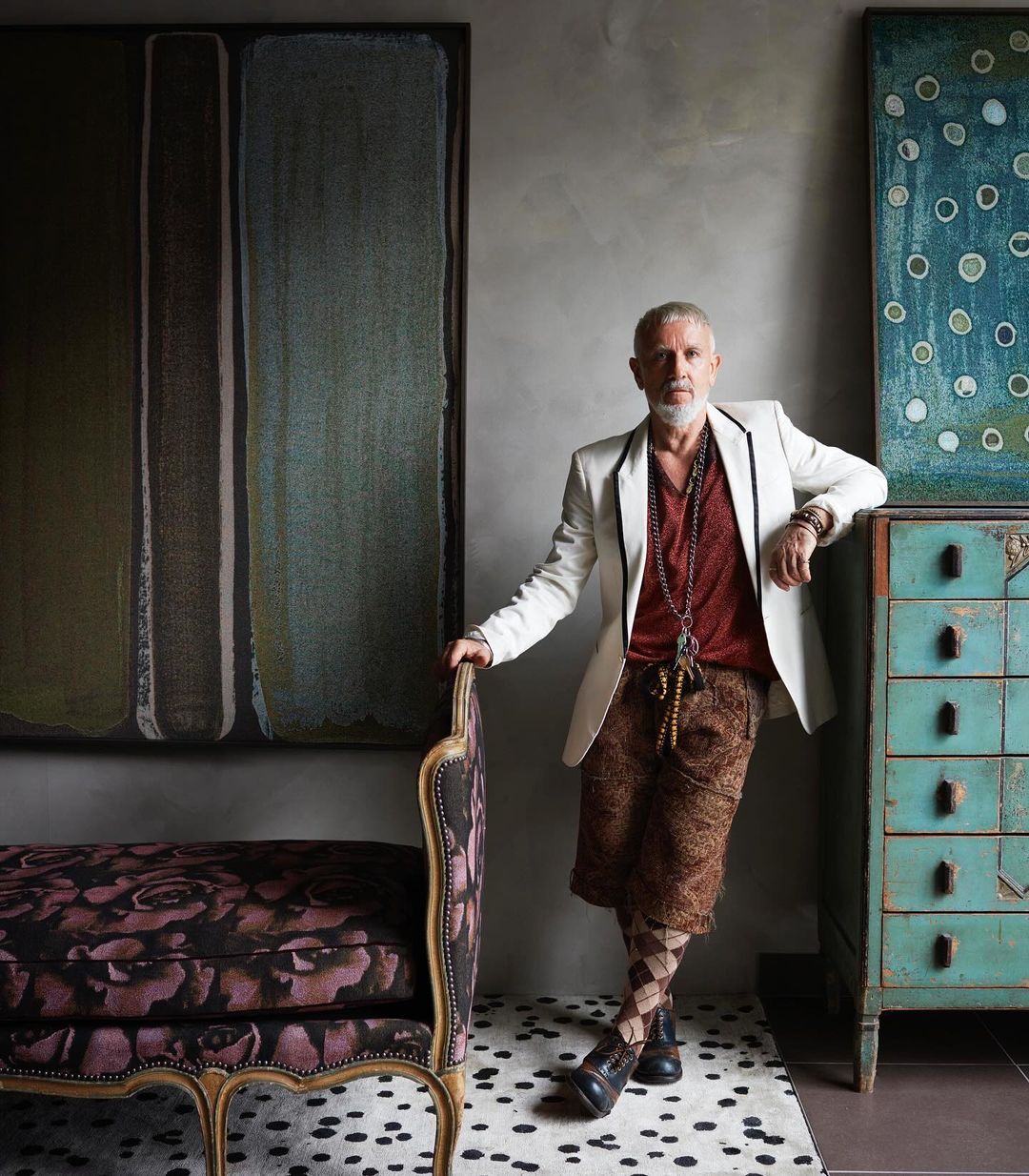 Stylish mature man in patterned shorts and jacket poses indoors with decorative furniture, art, carpet, and grey wall backdrop.