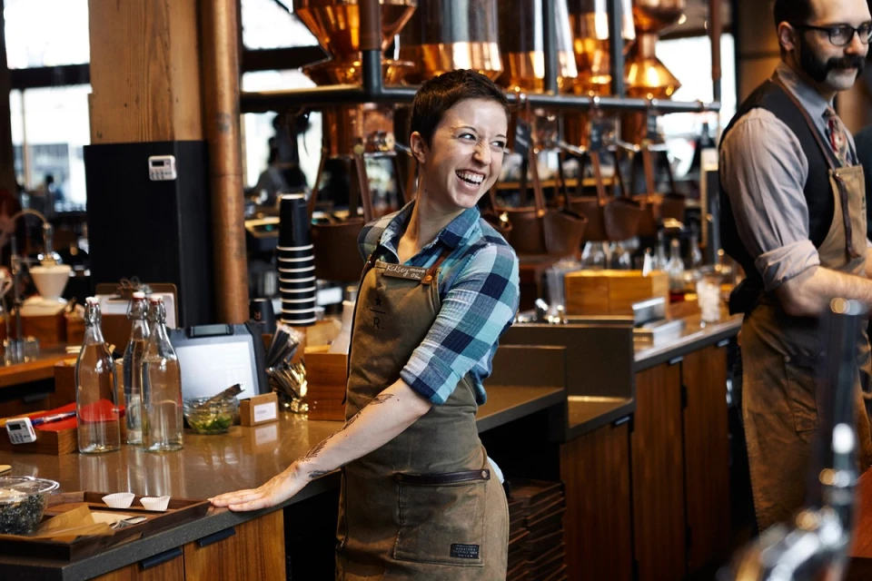 Woman smiles in a coffee shop, wearing an apron behind the counter, near coffee brewing equipment and beverage preparation supplies.