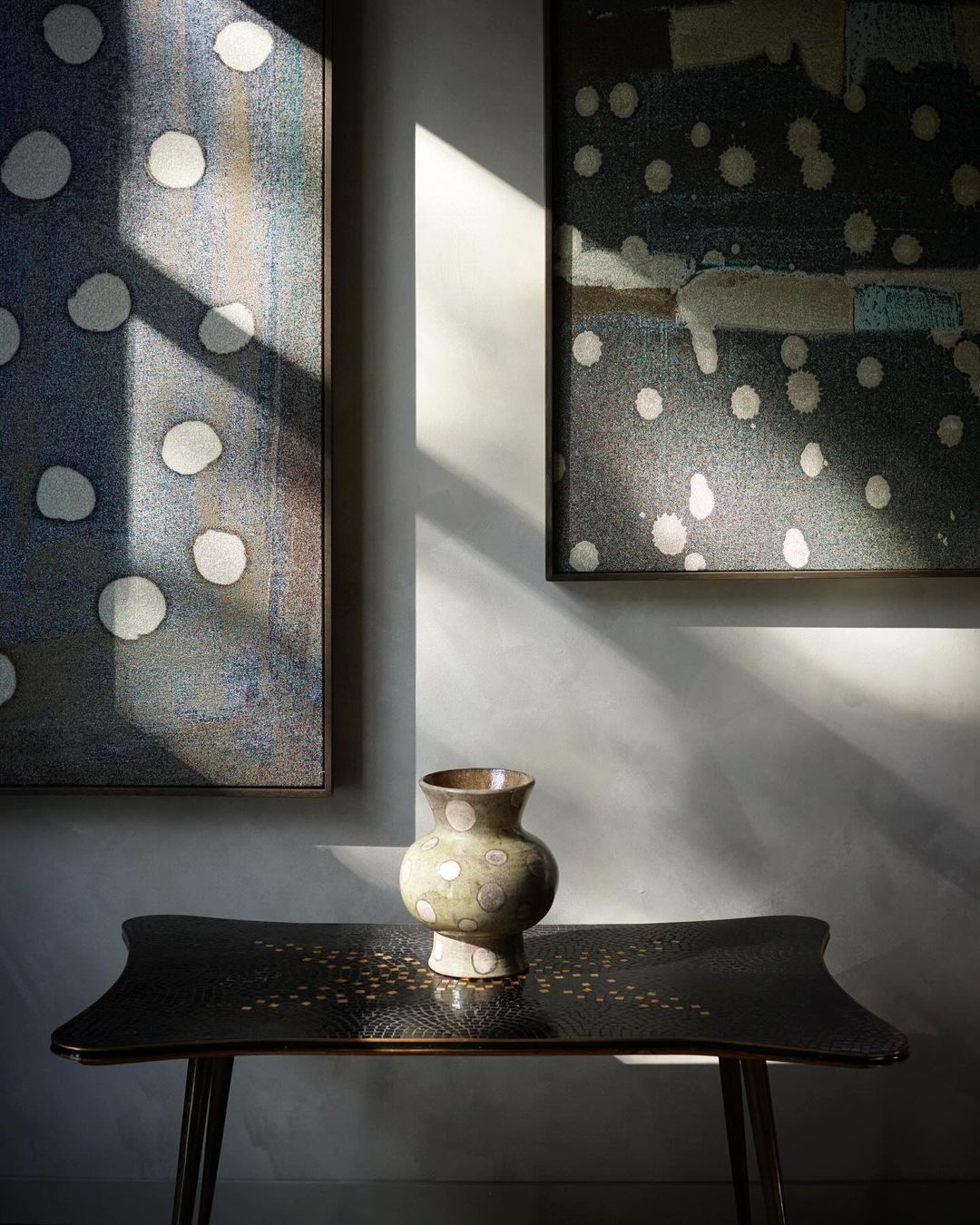 Still life featuring a decorative vase on a black table, framed by two abstract art pieces and natural light casting shadows on the wall.