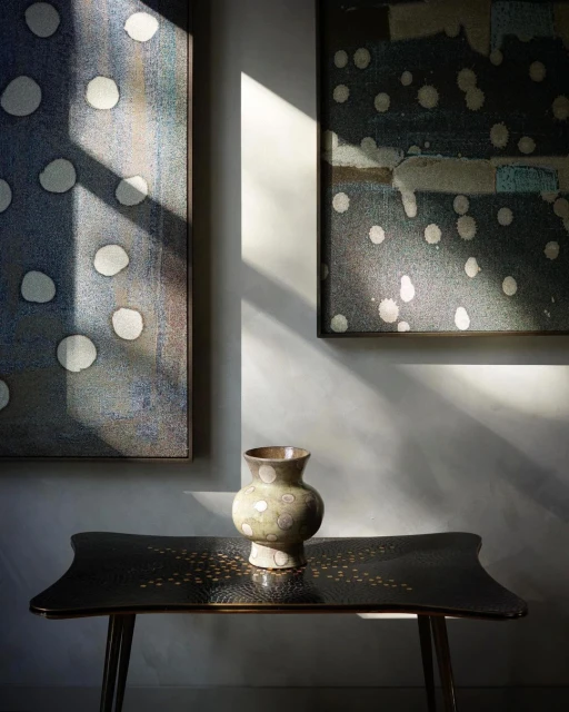 Still life featuring a decorative vase on a black table, framed by two abstract art pieces and natural light casting shadows on the wall.