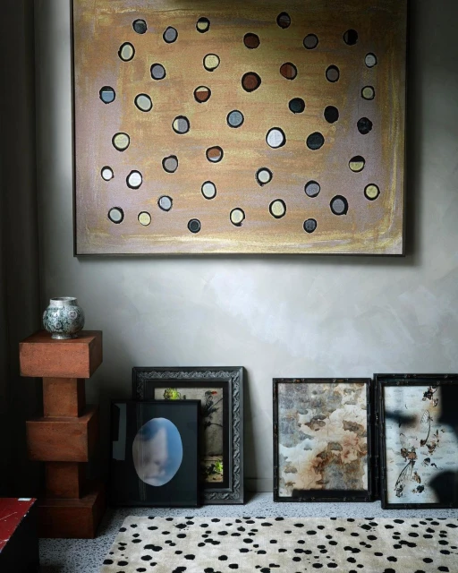 Interior with modern art: framed spot-patterned artwork, stacked table, vase, framed photos, and spotted rug on a neutral wall.