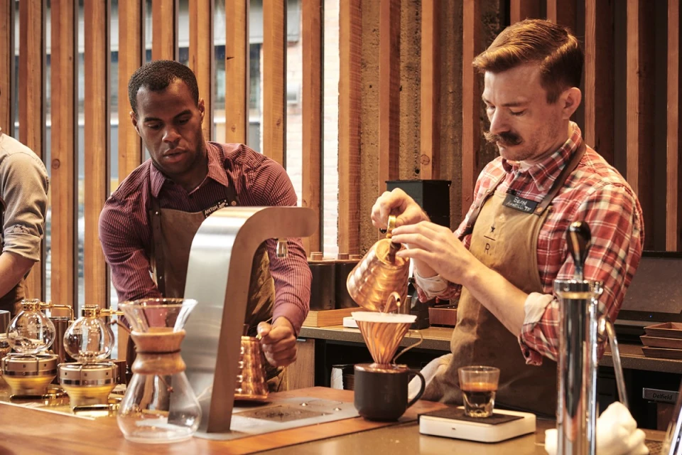Two baristas prepare coffee at a counter with professional equipment, wearing aprons in a warmly lit coffee shop setting.