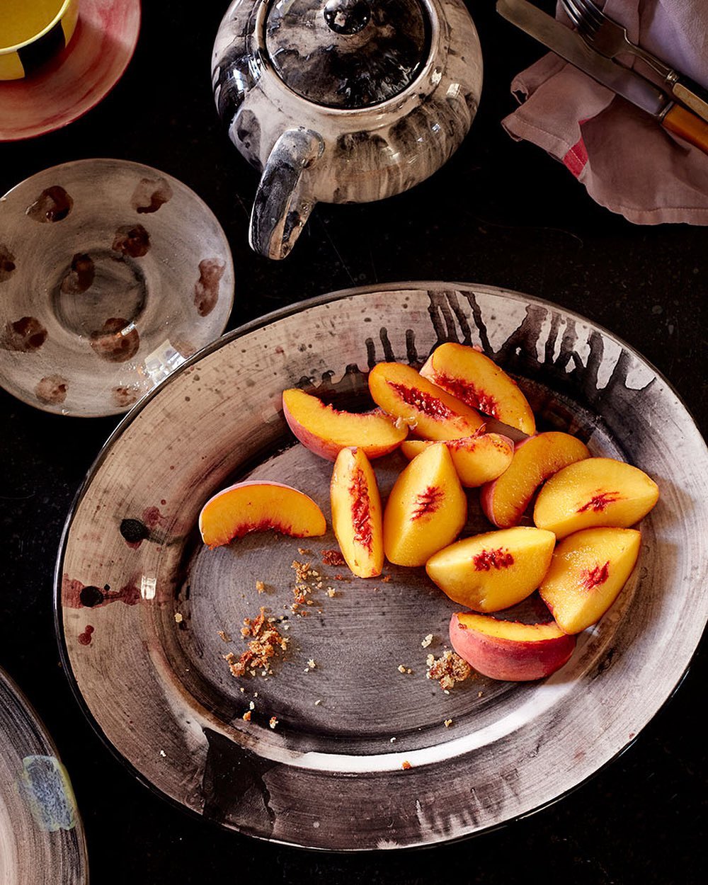 Overhead shot of sliced peaches on a painted plate, alongside a teapot, teacup, and silverware on a dark surface.