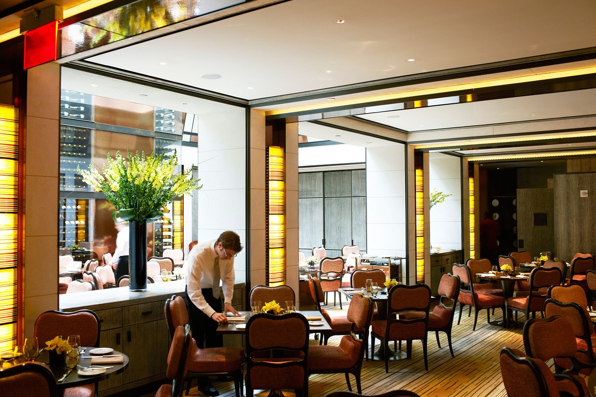 A waiter in a white shirt and tie sets a table inside an upscale restaurant with modern lighting and floral arrangements.
