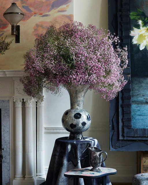 Close-up of a tall, silver vase holding a large bouquet of pink baby's breath flowers, set indoors with decorative wall elements.