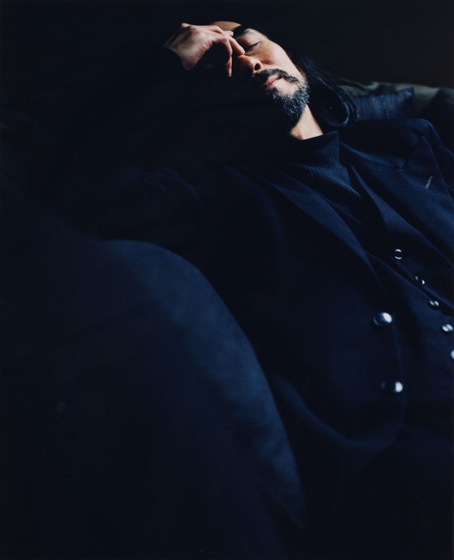 An adult male with a beard and long dark hair, wearing a black blazer, photographed indoors in dark, moody lighting.