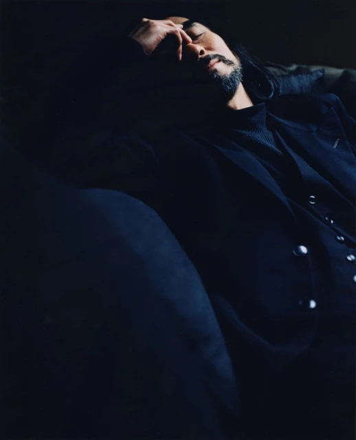 An adult male with a beard and long dark hair, wearing a black blazer, photographed indoors in dark, moody lighting.