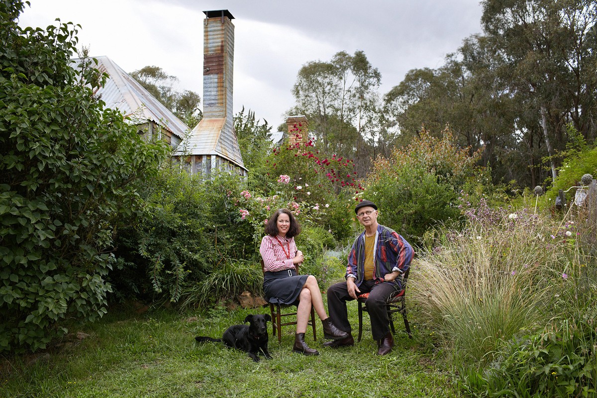 Adult couple and a black dog sitting in a lush green garden with a rustic house featuring a tall metal chimney and diverse plants.