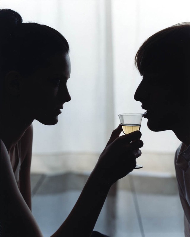 Silhouette of two people facing each other, one drinking white wine from a clear glass in a brightly lit indoor setting.