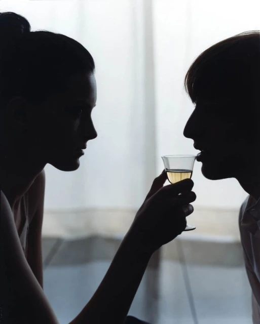 Silhouette of two people facing each other, one drinking white wine from a clear glass in a brightly lit indoor setting.