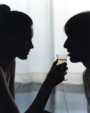 Silhouette of two people facing each other, one drinking white wine from a clear glass in a brightly lit indoor setting.
