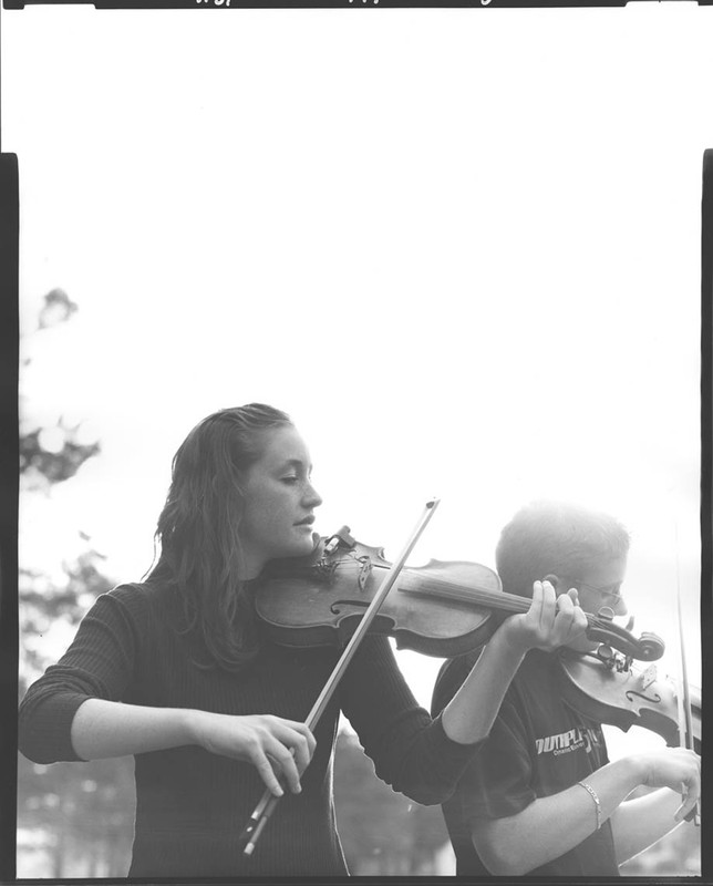Two people are playing violins outdoors. One is an adult with long hair, and the other person is a child wearing a graphic t-shirt.