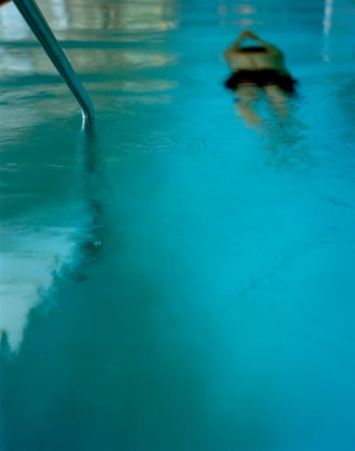 Person swimming in a clear, turquoise pool with sunlight and visible ladder for pool access in a vertical, indoor shot.
