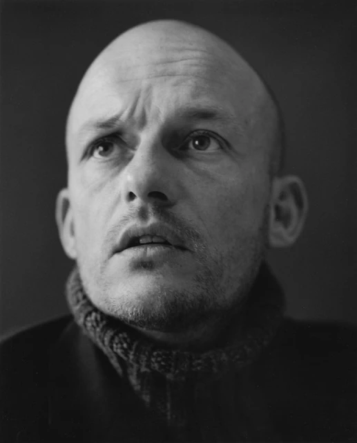 Portrait of an adult man with a bald head wearing a sweater. The black and white portrait is shot at eye level.