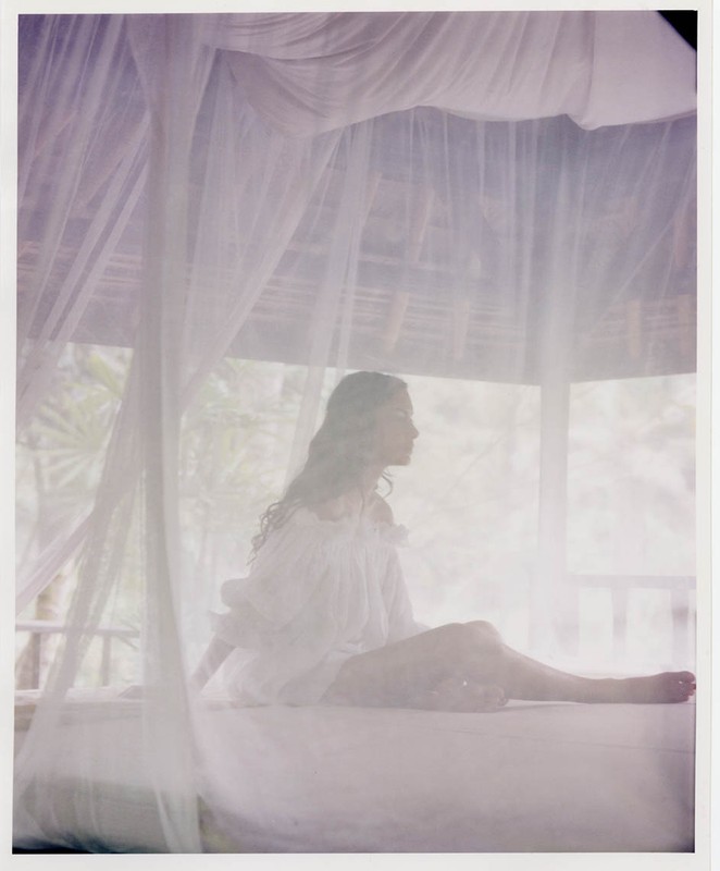 Woman in white dress sits on bed with mosquito netting in tropical outdoor cabana, enjoying soft light and relaxed ambiance.