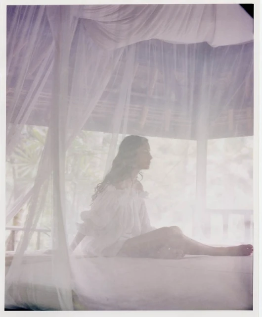 Woman in white dress sits on bed with mosquito netting in tropical outdoor cabana, enjoying soft light and relaxed ambiance.