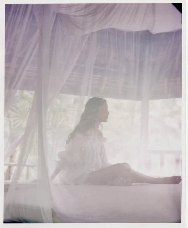 Woman in white dress sits on bed with mosquito netting in tropical outdoor cabana, enjoying soft light and relaxed ambiance.