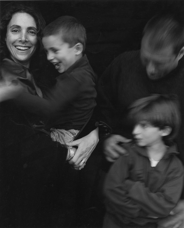 Monochrome family portrait with a smiling woman holding a child and another child in the foreground by Martyn Thompson