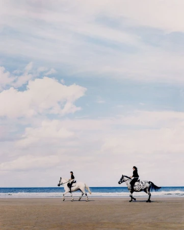 Two women ride horses on a sandy beach with the ocean waves crashing and a cloudy sky above in this outdoor nature scene.