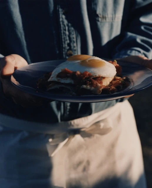 Person holding a plate with fried eggs on top of a dish with sauce. The image has a warm, inviting feel with natural light.