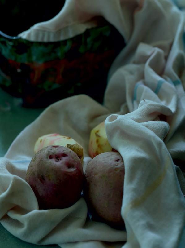 Close-up still life of whole and peeled red potatoes with a white cloth, and a glimpse of a black bowl in dim, moody lighting.
